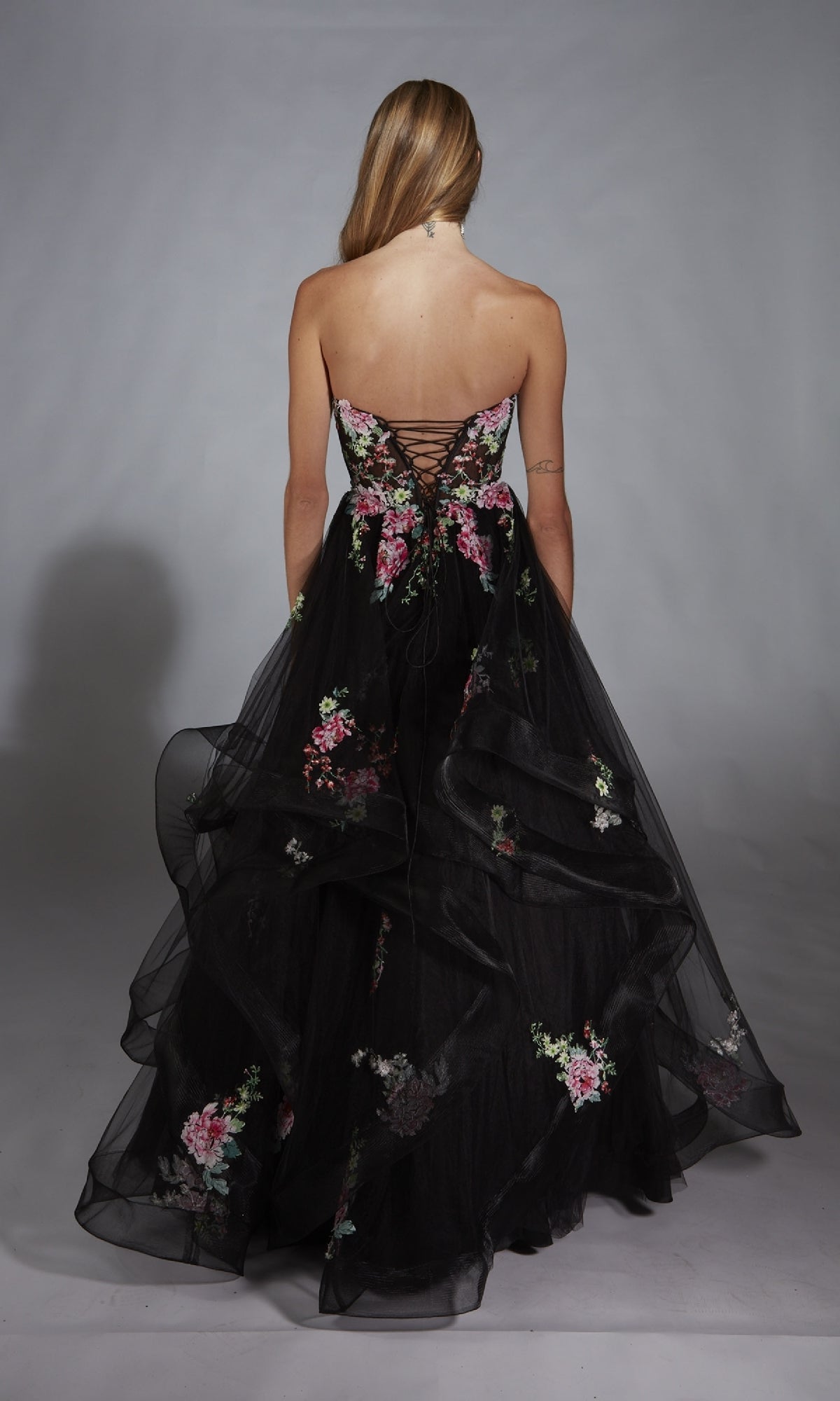 A woman with long hair models the Alyce 61914 long prom dress, a strapless ball gown with floral embroidery and a lace-up back, posed against a plain gray background.